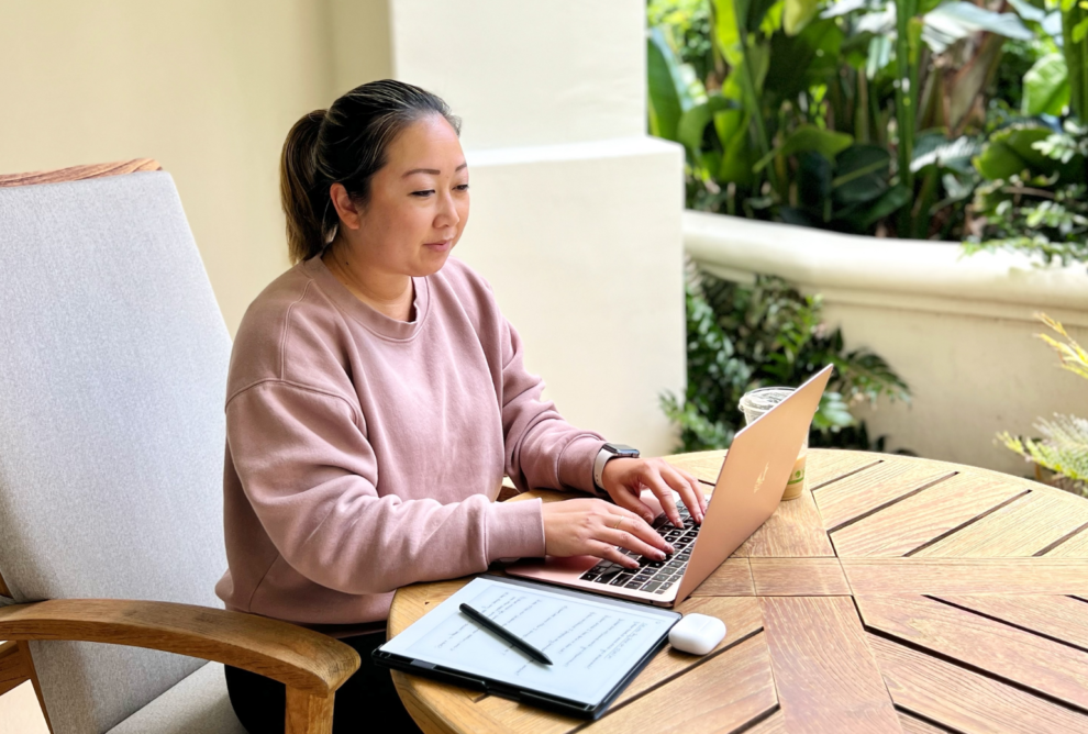 Asian-American woman working on a laptop with tropical plants on the upper right corner